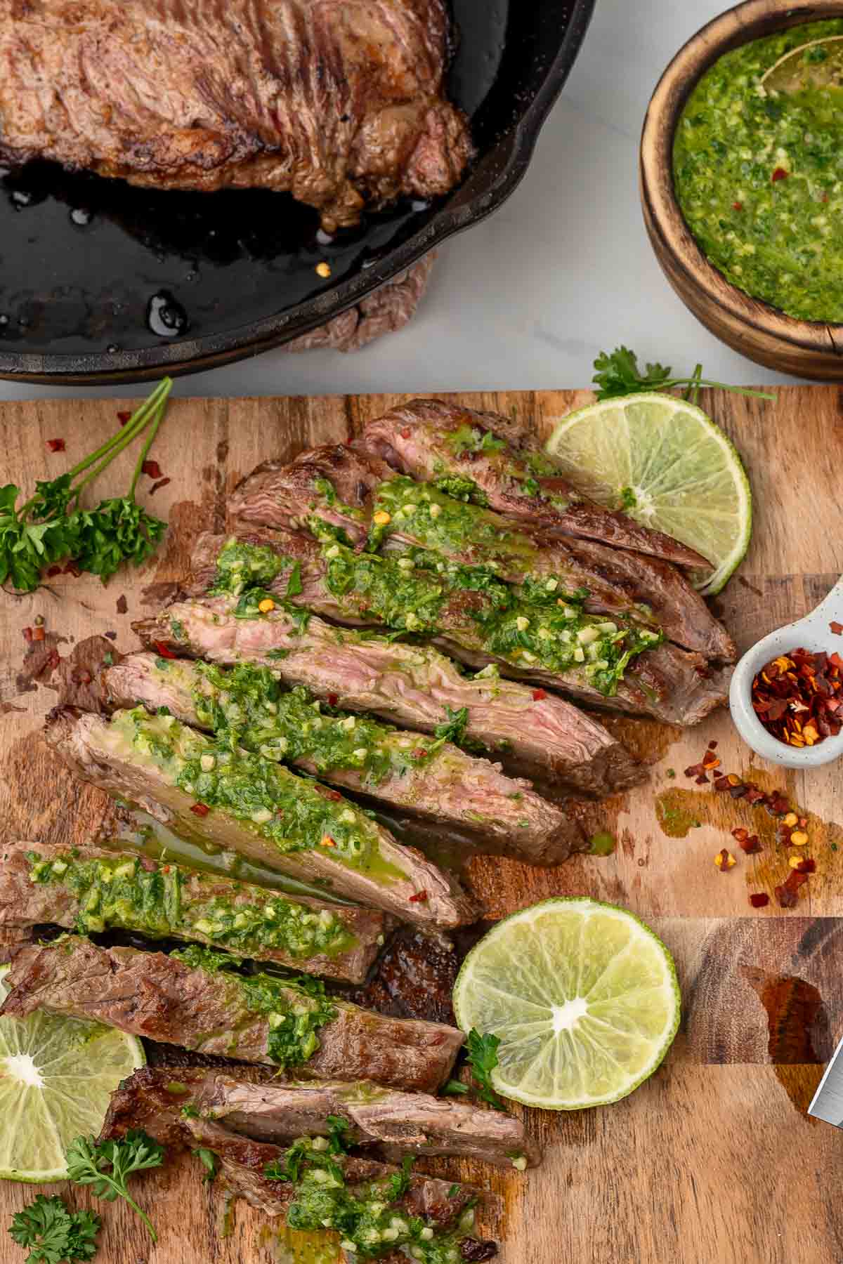 Sliced pan-seared skirt steak topped with green chimichurri sauce on a wooden board, garnished with lime slices, parsley, and a bowl of sauce.