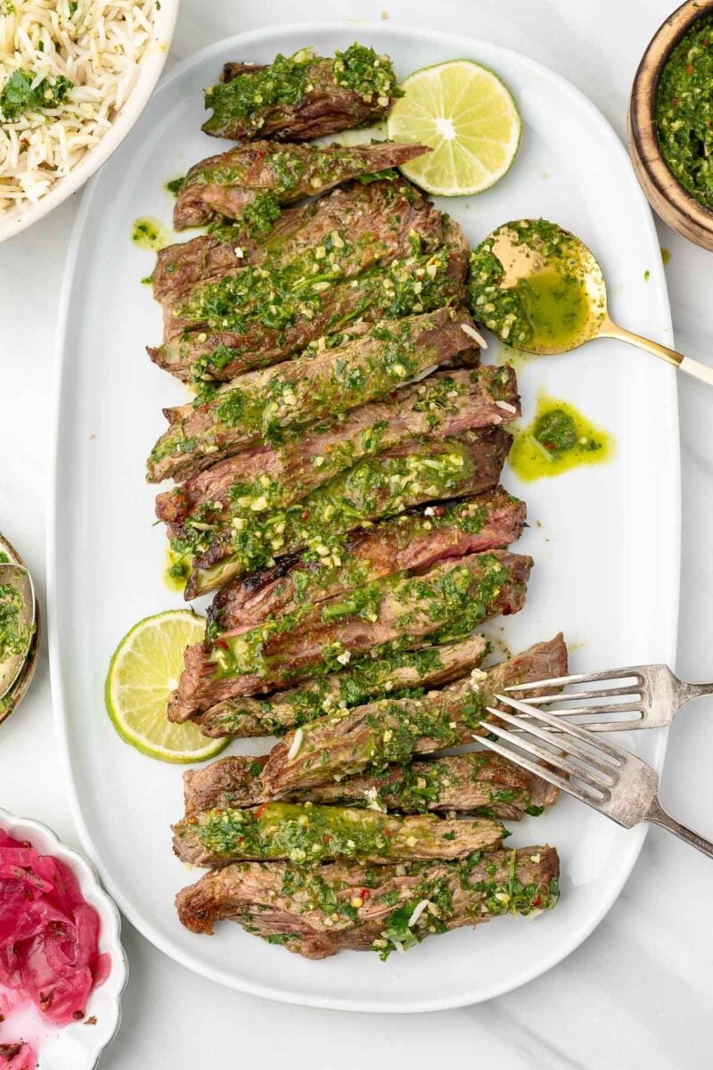 Sliced skirt steak topped with green chimichurri sauce, served on a white platter with lime slices and a fork.