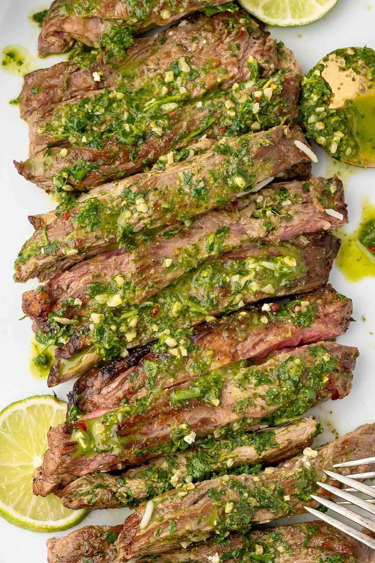 Sliced skirt steak topped with green chimichurri sauce, garnished with lime wedges on a white plate.