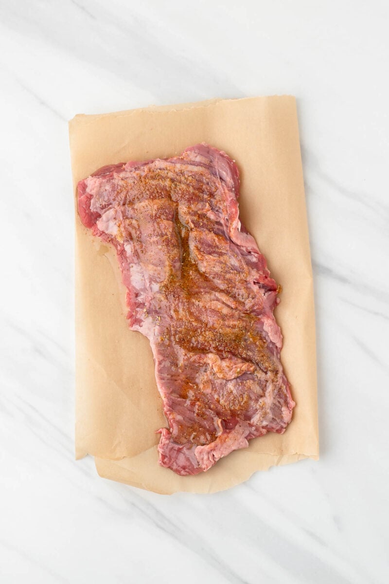 Raw skirt steak seasoned with spices, placed on a sheet of brown parchment paper on a white marble surface.