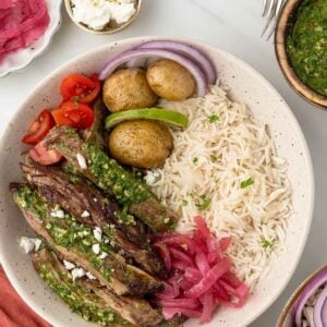 A bowl with sliced skirt steak topped with green sauce, rice, roasted potatoes, cherry tomatoes, red onion, pickled onions, sliced lime, and a sprinkle of feta cheese.