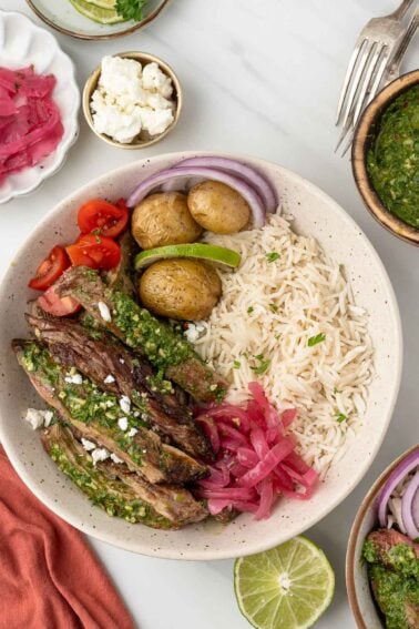 A bowl with sliced skirt steak topped with green sauce, rice, roasted potatoes, cherry tomatoes, red onion, pickled onions, sliced lime, and a sprinkle of feta cheese.