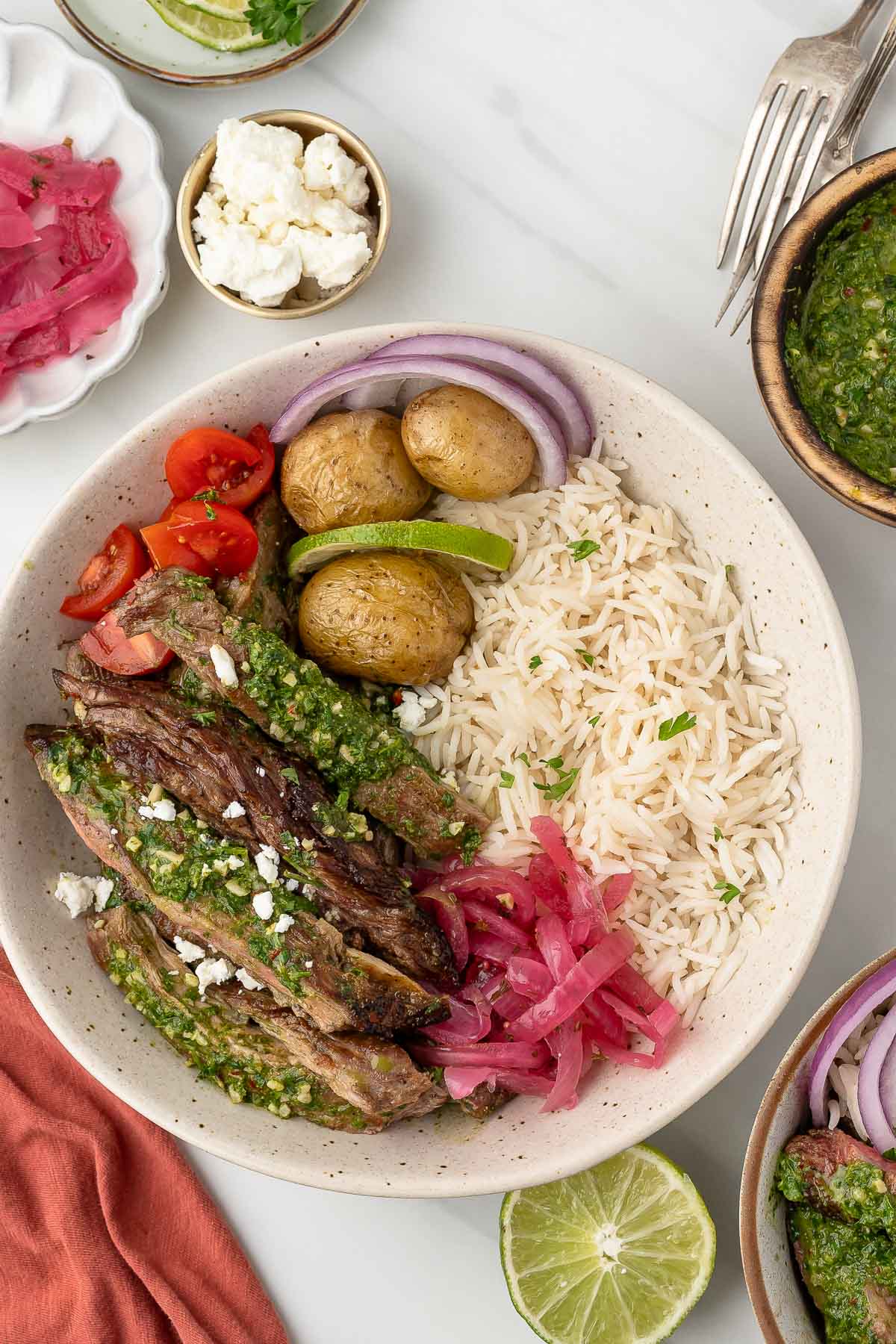 A bowl with sliced skirt steak topped with green sauce, rice, roasted potatoes, cherry tomatoes, red onion, pickled onions, sliced lime, and a sprinkle of feta cheese.