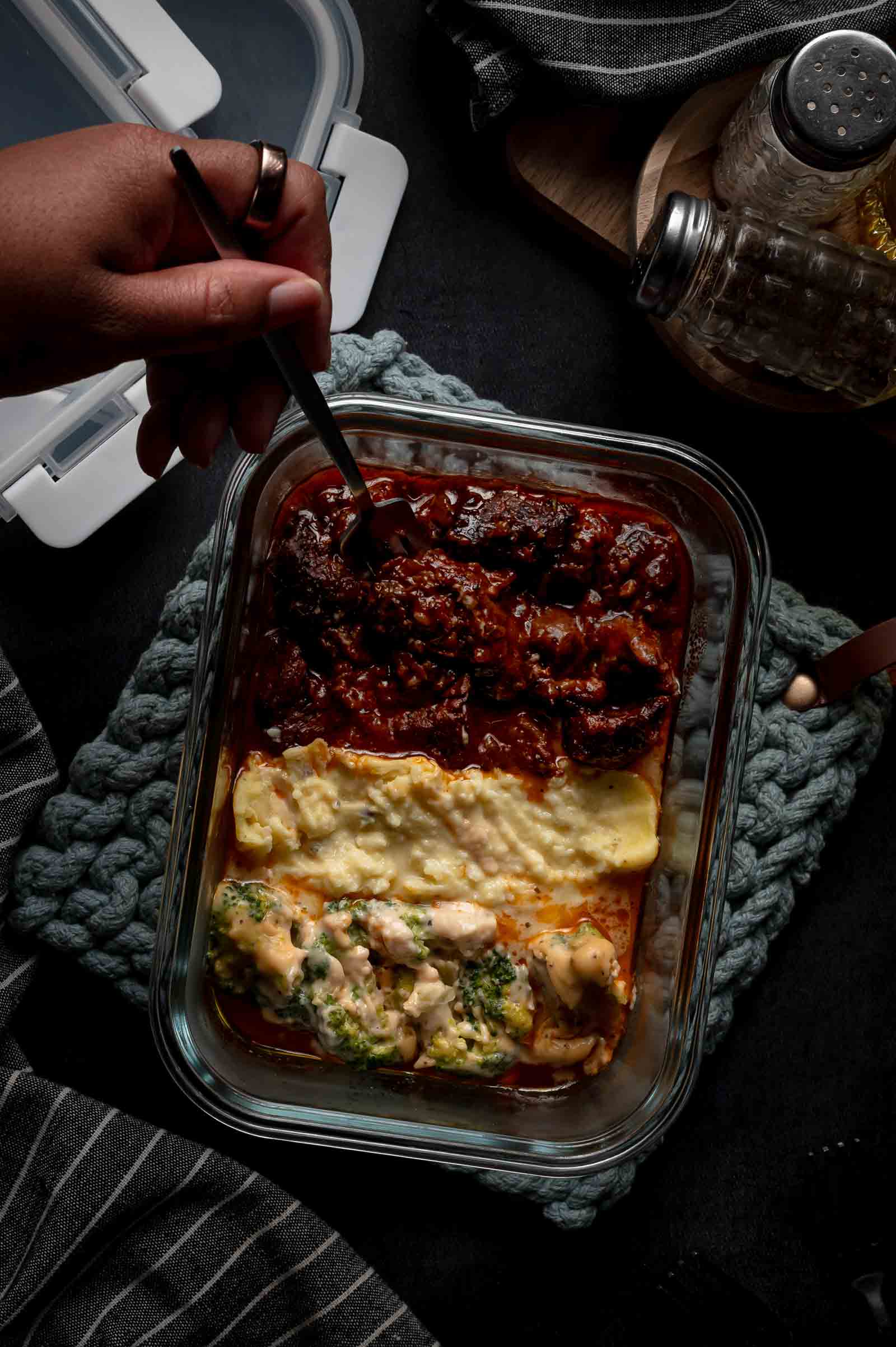 A hand holds a fork over a glass container with beef tips, mashed potatoes and broccoli, on a dark background with a pepper shaker nearby.