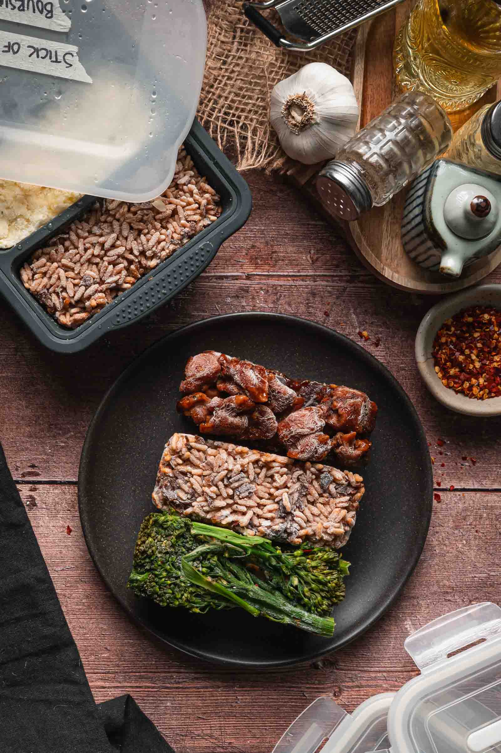 A black plate with frozen meal portions of bourbon chicken, brown rice, and garlic broccolini; nearby more silicon freezer molds with frozen meals.