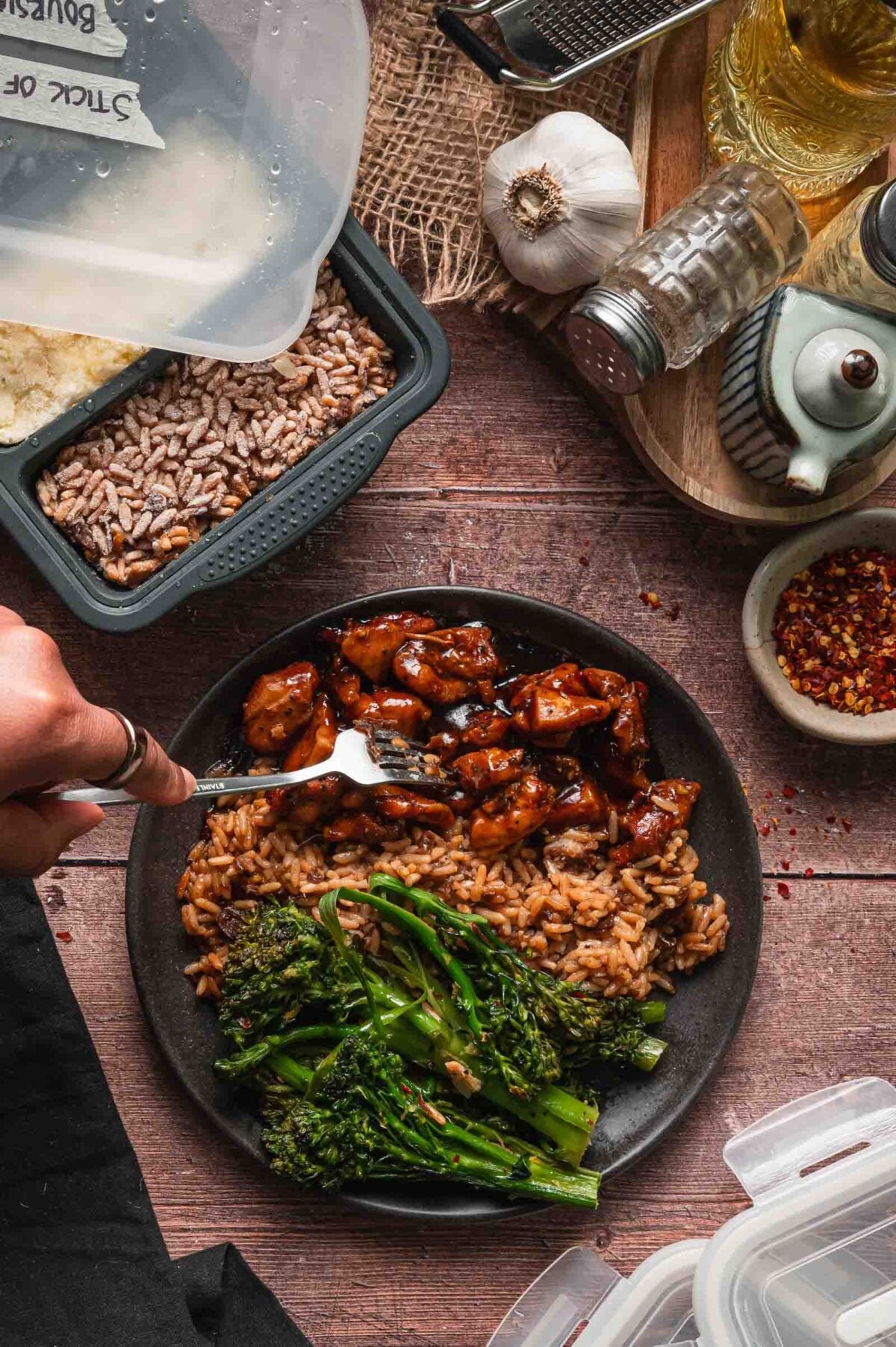 A plate with cooked stick of butter rice, glazed bourbon chicken pieces, and lemony garlic broccolini, with a hand holding a fork; meal prep containers and seasonings are visible nearby.