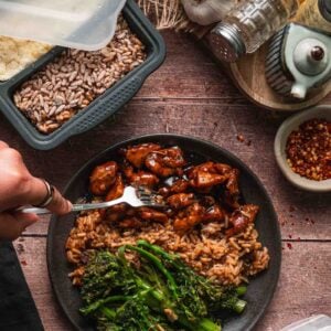 A plate with cooked stick of butter rice, glazed bourbon chicken pieces, and lemony garlic broccolini, with a hand holding a fork; meal prep containers and seasonings are visible nearby.
