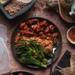 A plate of bourbon chicken with stick of butter rice and lemon garlic broccolini, surrounded by meal prep containers, spices, and kitchen utensils on a rustic wooden surface.