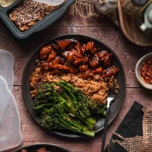 A plate of bourbon chicken with stick of butter rice and lemon garlic broccolini, surrounded by meal prep containers, spices, and kitchen utensils on a rustic wooden surface.