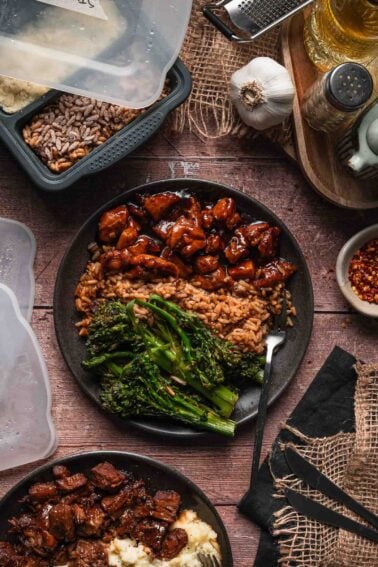 A plate of bourbon chicken with stick of butter rice and lemon garlic broccolini, surrounded by meal prep containers, spices, and kitchen utensils on a rustic wooden surface.