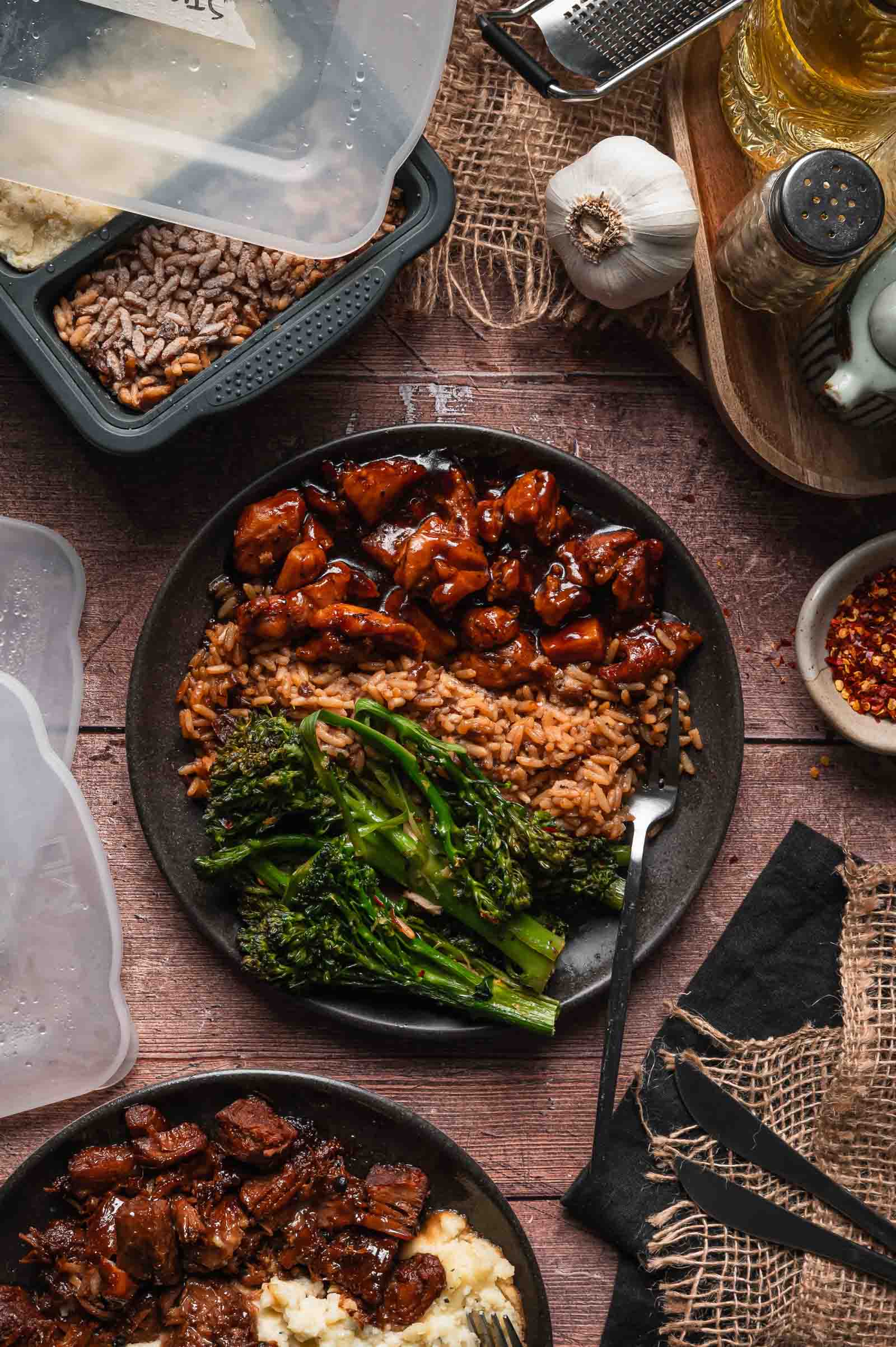 A plate of bourbon chicken with stick of butter rice and lemon garlic broccolini, surrounded by meal prep containers, spices, and kitchen utensils on a rustic wooden surface.
