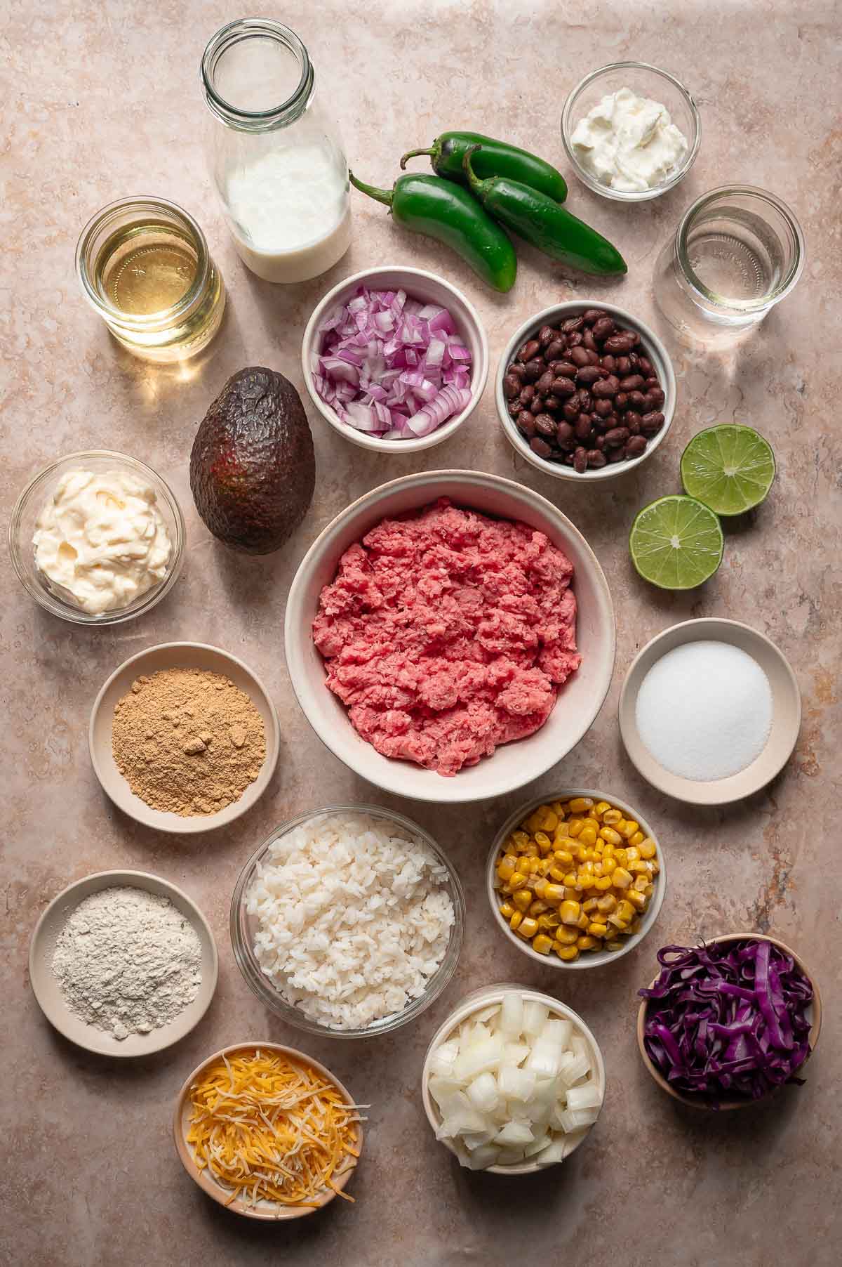 Overhead view of ground beef, black beans, avocado, corn, chopped onions, jalapeรฑos, shredded cheese, lime halves, spices, flour, oil, and dairy in small bowls on a countertop.