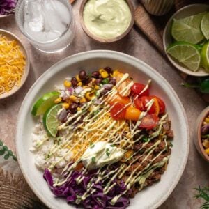 A burrito bowl with rice, shredded beef, corn, black beans, tomatoes, cabbage, cheese, lime, and sauce, served with various toppings and drinks on a table.