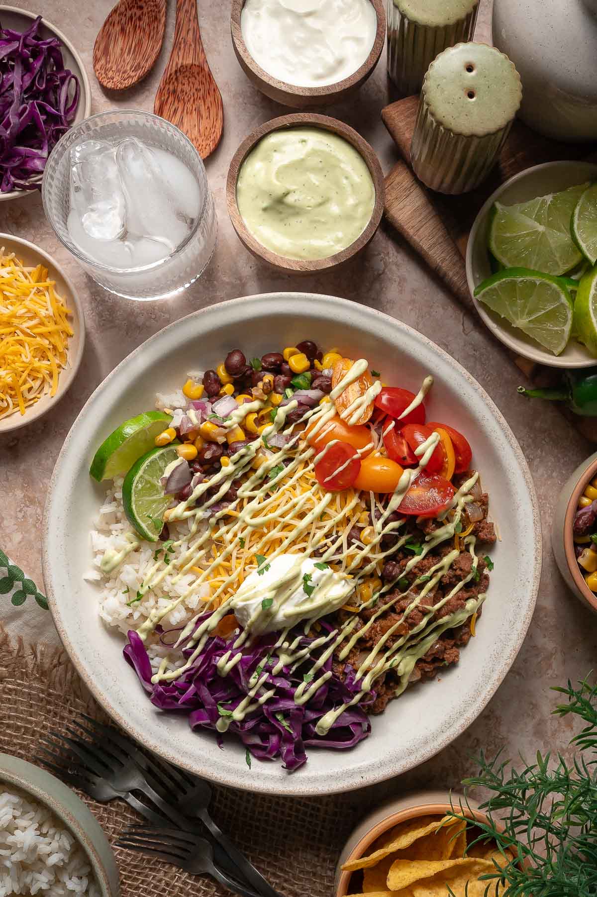 A burrito bowl with rice, shredded beef, corn, black beans, tomatoes, cabbage, cheese, lime, and sauce, served with various toppings and drinks on a table.