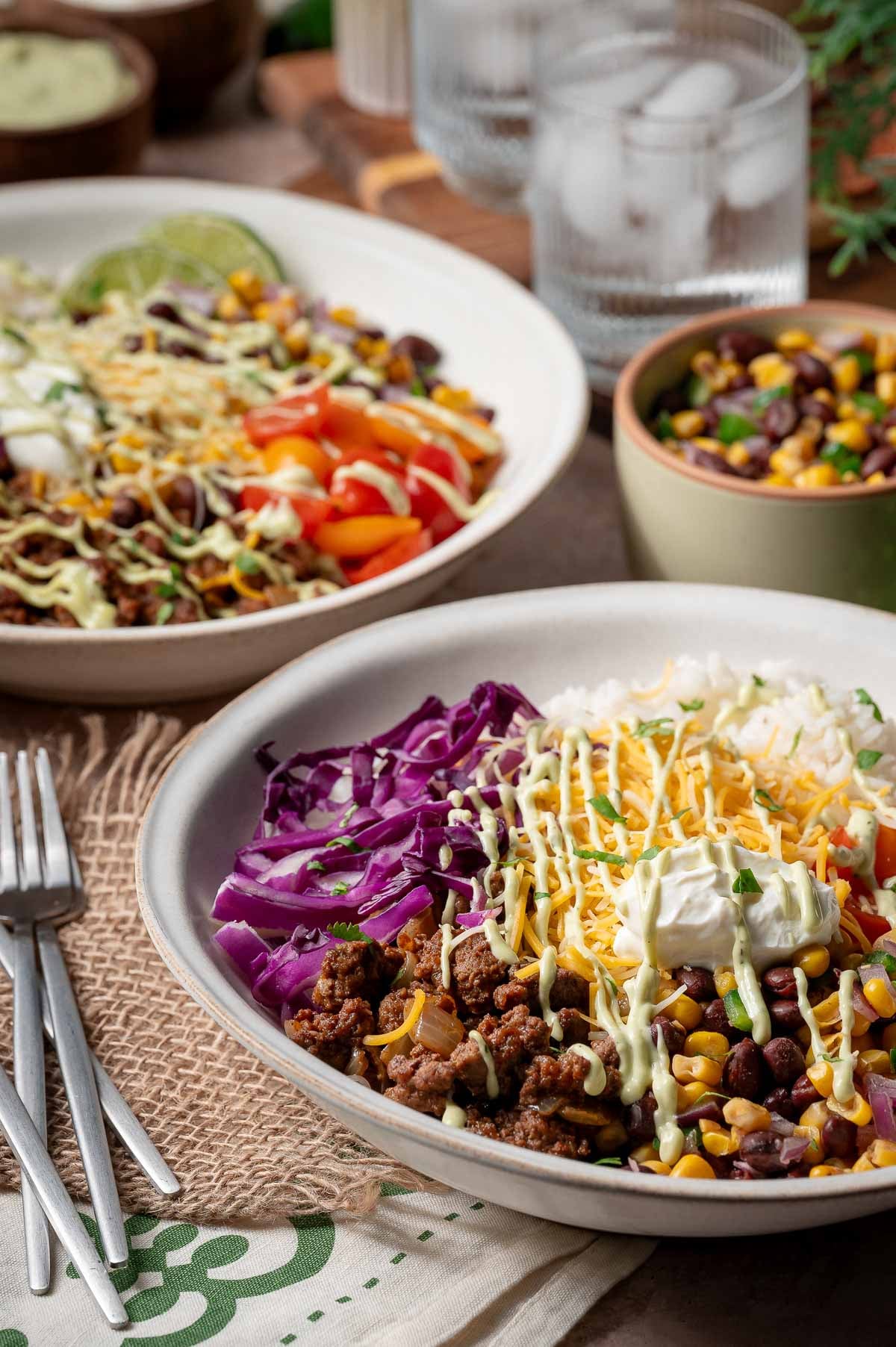 Two burrito bowls filled with rice, ground beef, shredded cheese, sour cream, red cabbage, corn, beans, and diced tomatoes, with forks and a small bowl of bean and corn salad on the side.