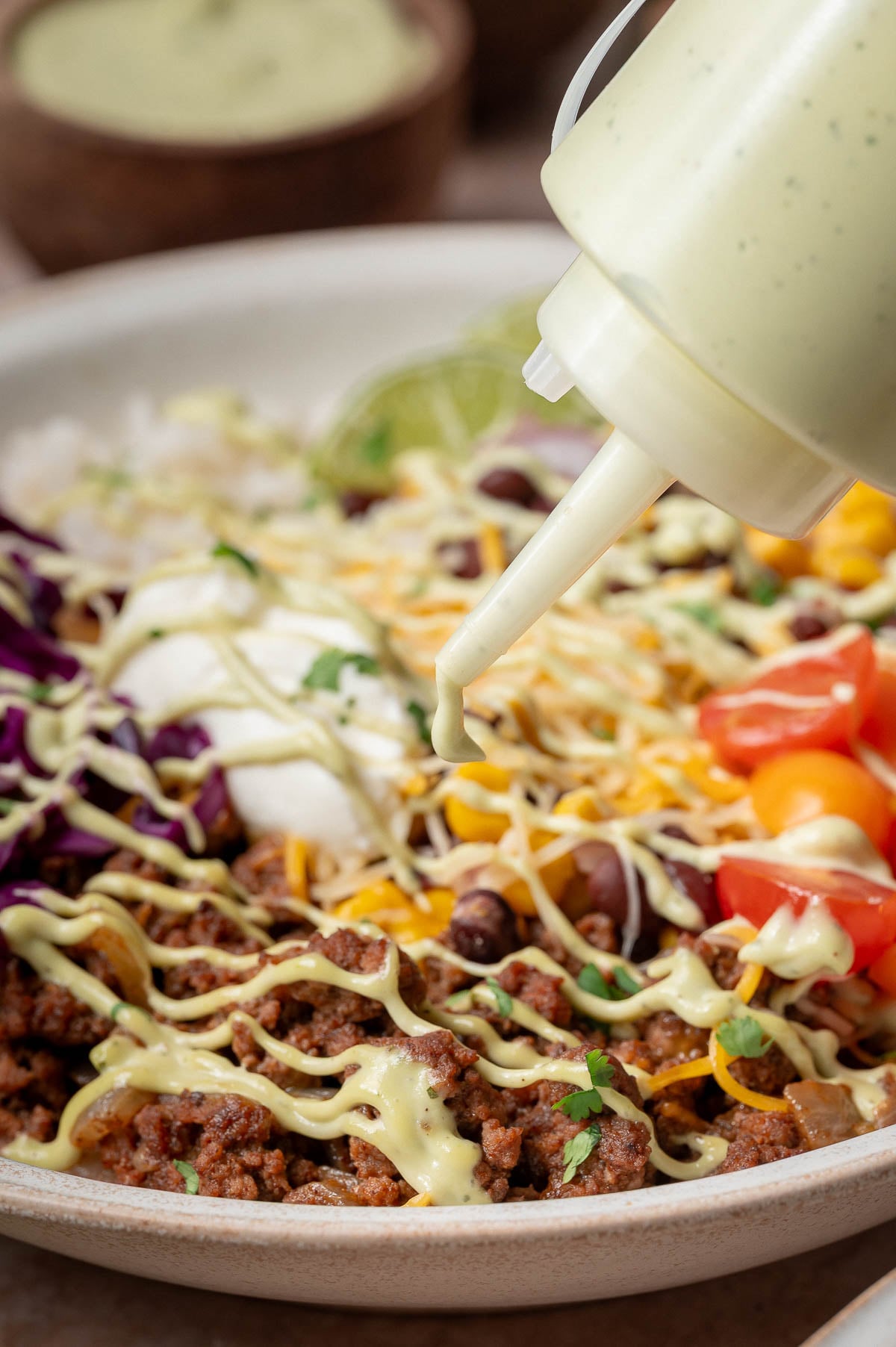 A close-up of a burrito bowl with ground beef, beans, cheese, vegetables, and avocado sauce being drizzled from a squeeze bottle.