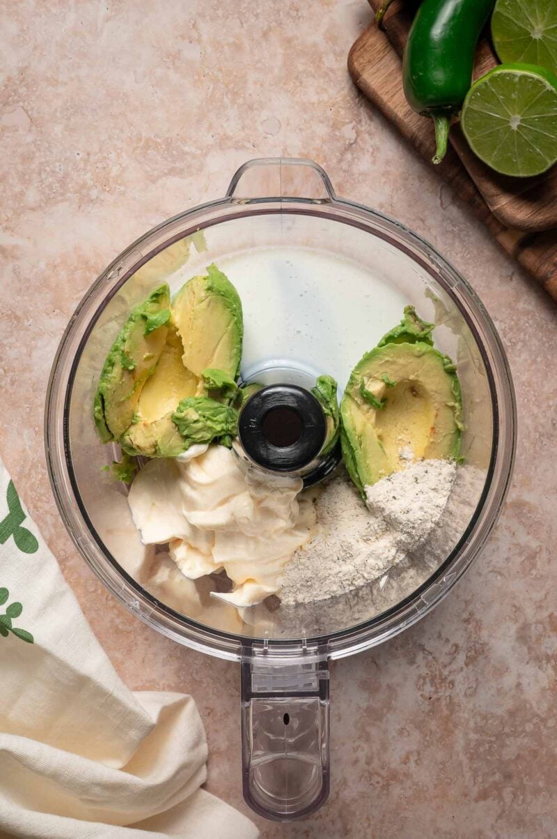 A food processor bowl with avocado halves, mayonnaise, a powder ingredient, and a white liquid. Jalapeรฑo, lime halves, and a wooden board are visible in the background.