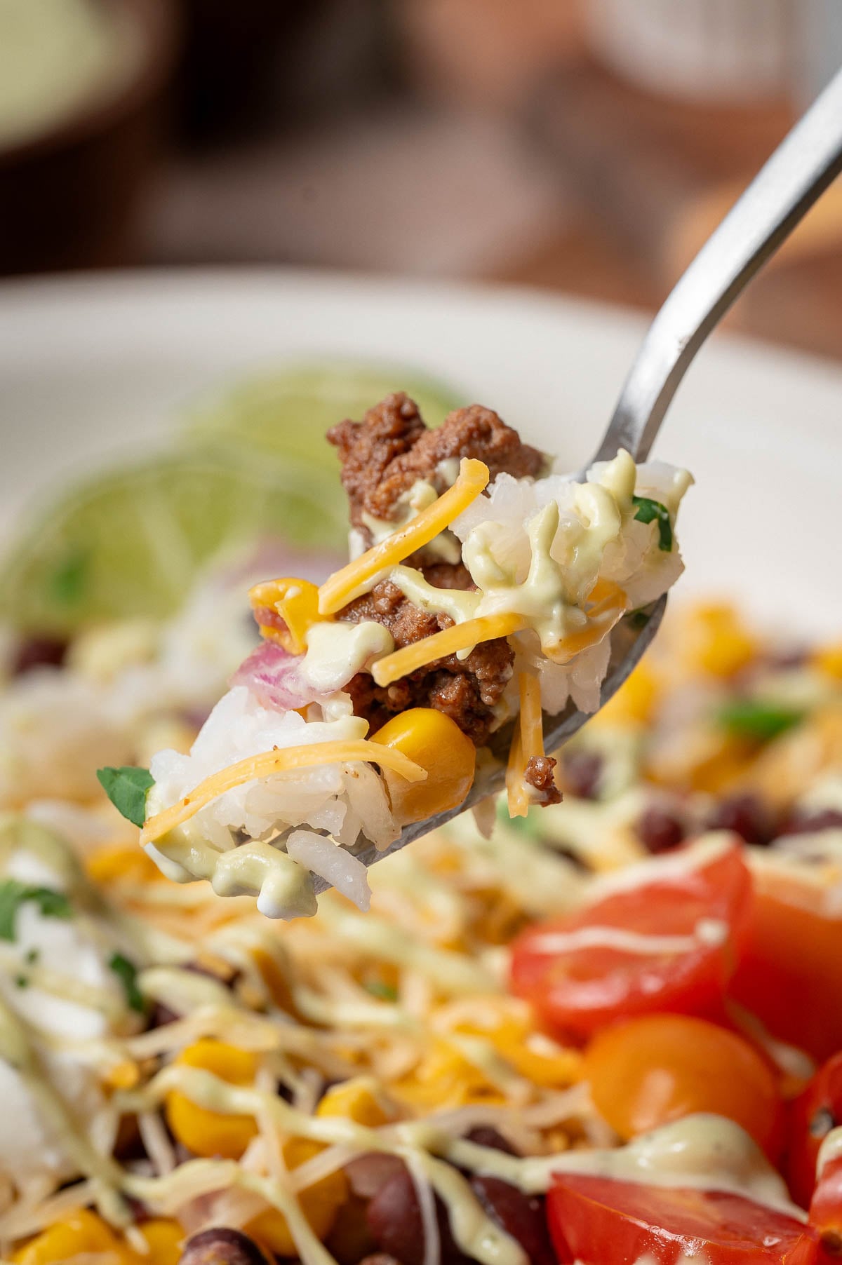 A fork holds a bite of rice, ground beef, corn, shredded cheese, and sauce above a burrito bowl, including black beans, tomatoes, and lime.