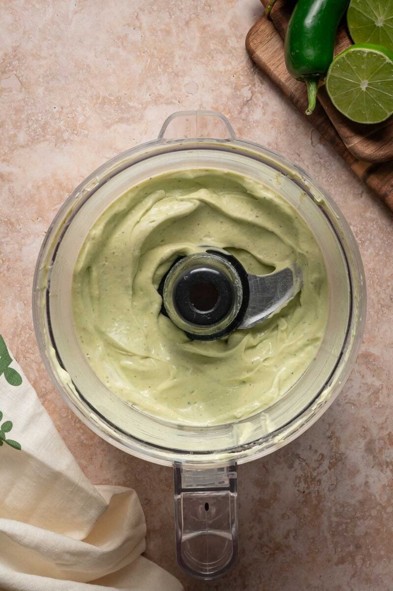 A food processor bowl filled with creamy green avocado sauce, with sliced limes and a jalapeรฑo on a cutting board in the background.