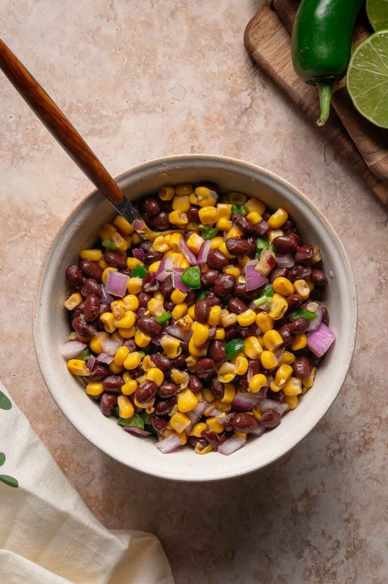 A bowl of corn and black bean salad with chopped red onion and jalapeรฑo, a spoon, and lime and jalapeรฑo on a cutting board in the background.