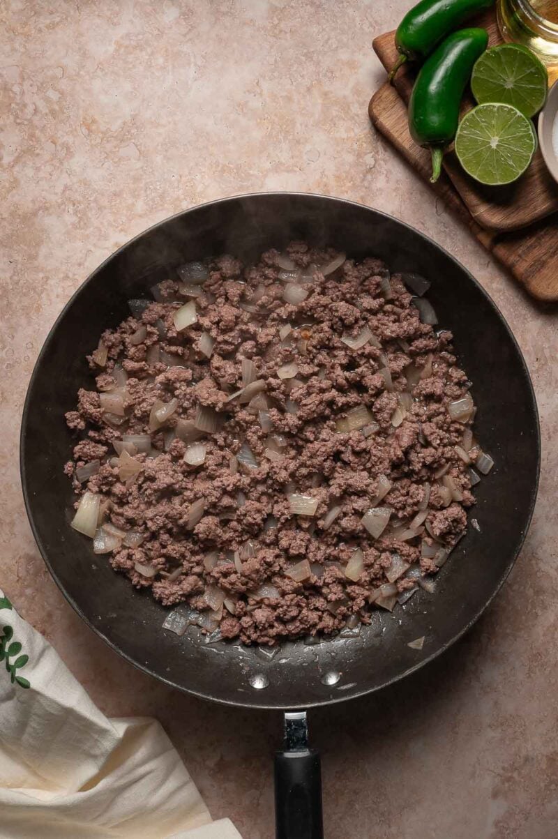 Ground beef and chopped onions cooking in a black skillet, with jalapeรฑos, lime, and a small bottle of oil on a wooden board nearby.