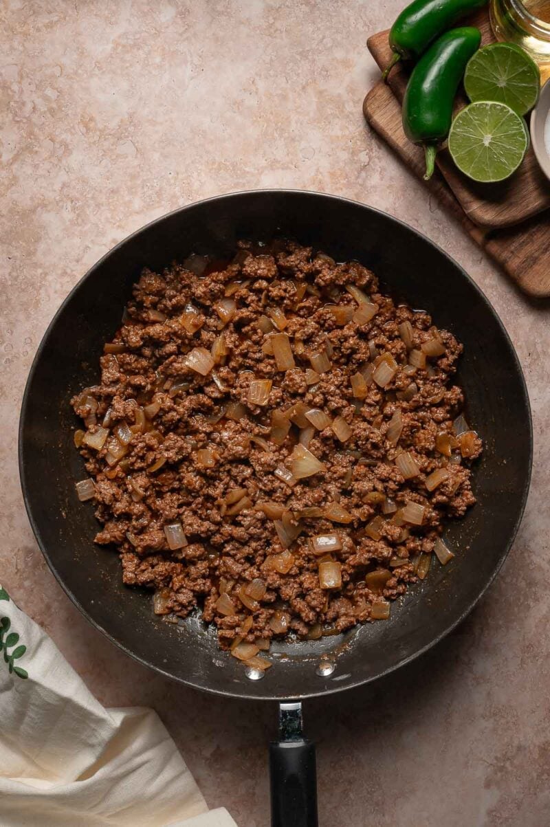 Taco spiced ground beef and diced onions cooked in a skillet, with jalapeรฑos, a halved lime, and a bowl of oil on a cutting board nearby.