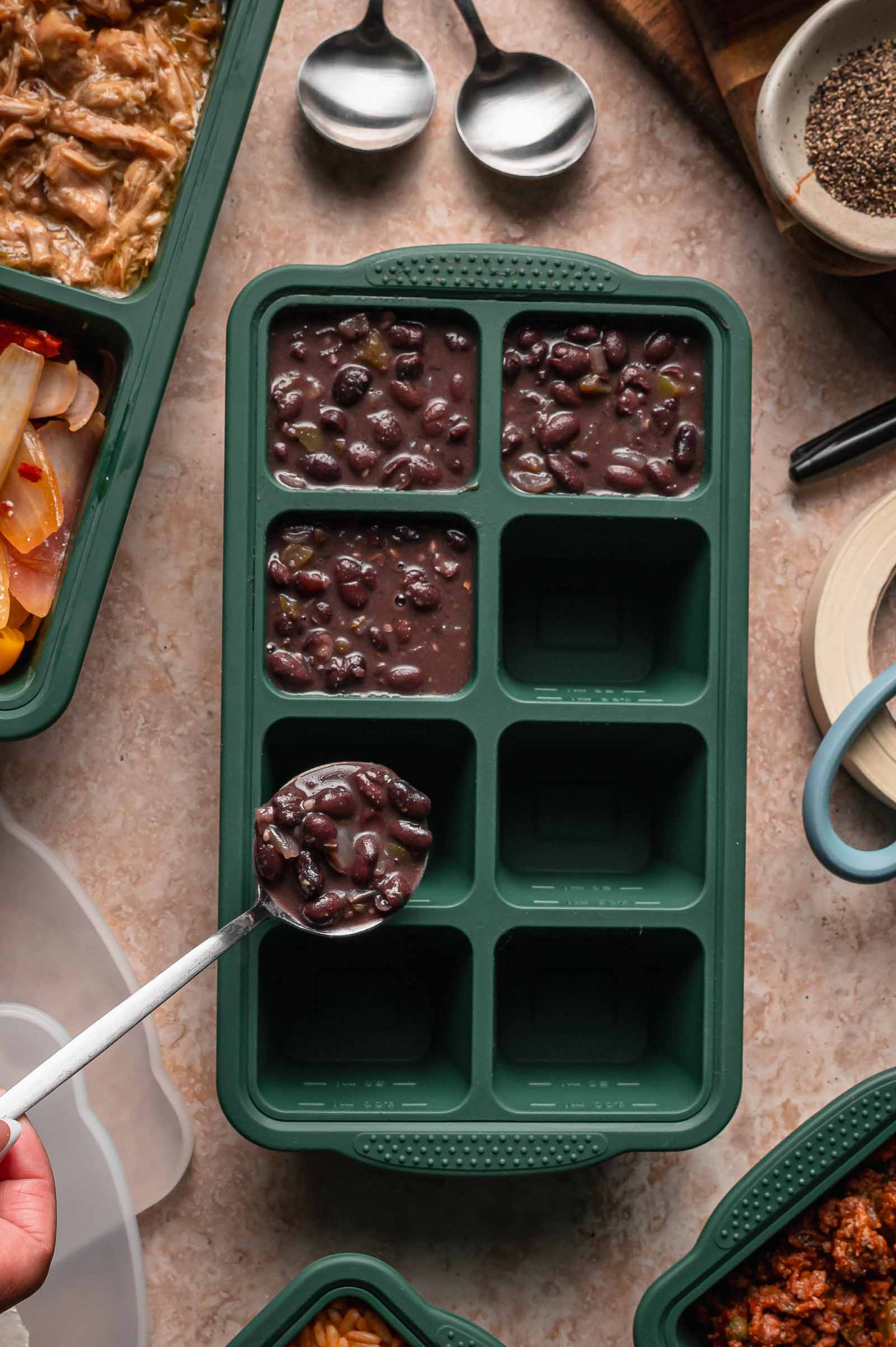 A hand ladles Cuban black beans into a green silicone freezer tray with six compartments, surrounded by food containers, spoons, and kitchen items on a countertop.