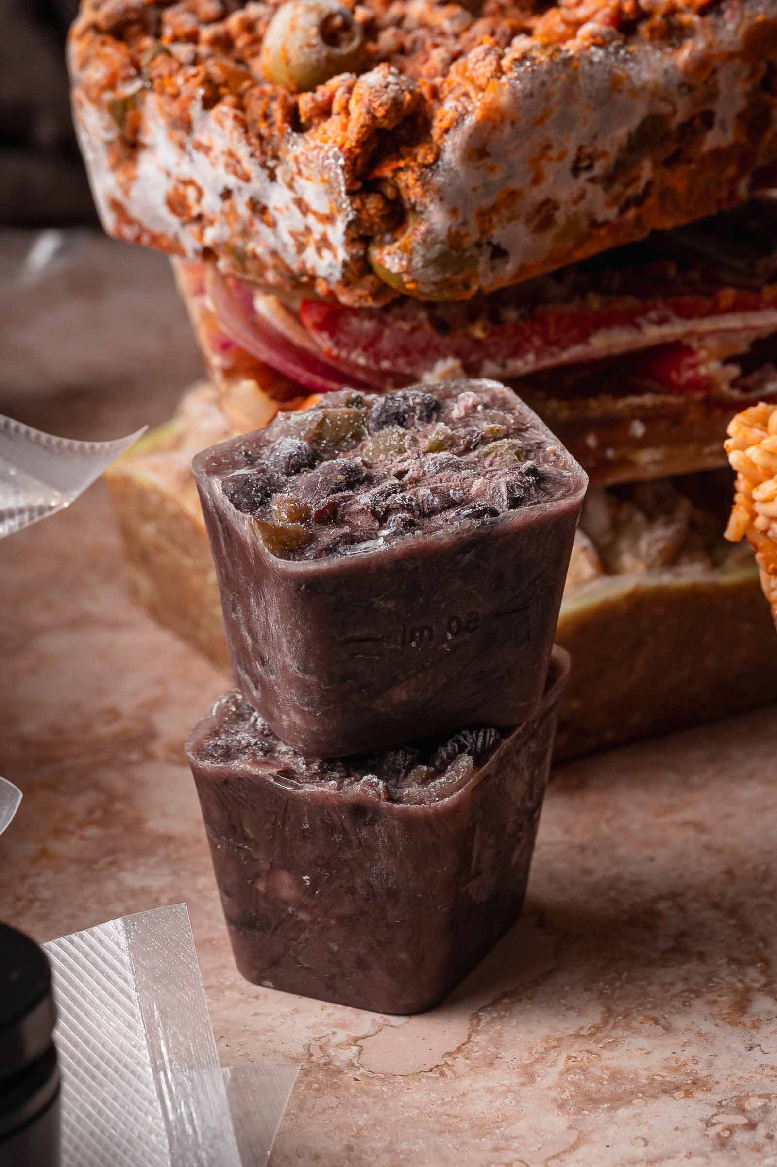 Two frozen cubes of cuban black beans, stacked on a countertop with more frozen meal prep cubes in the background.