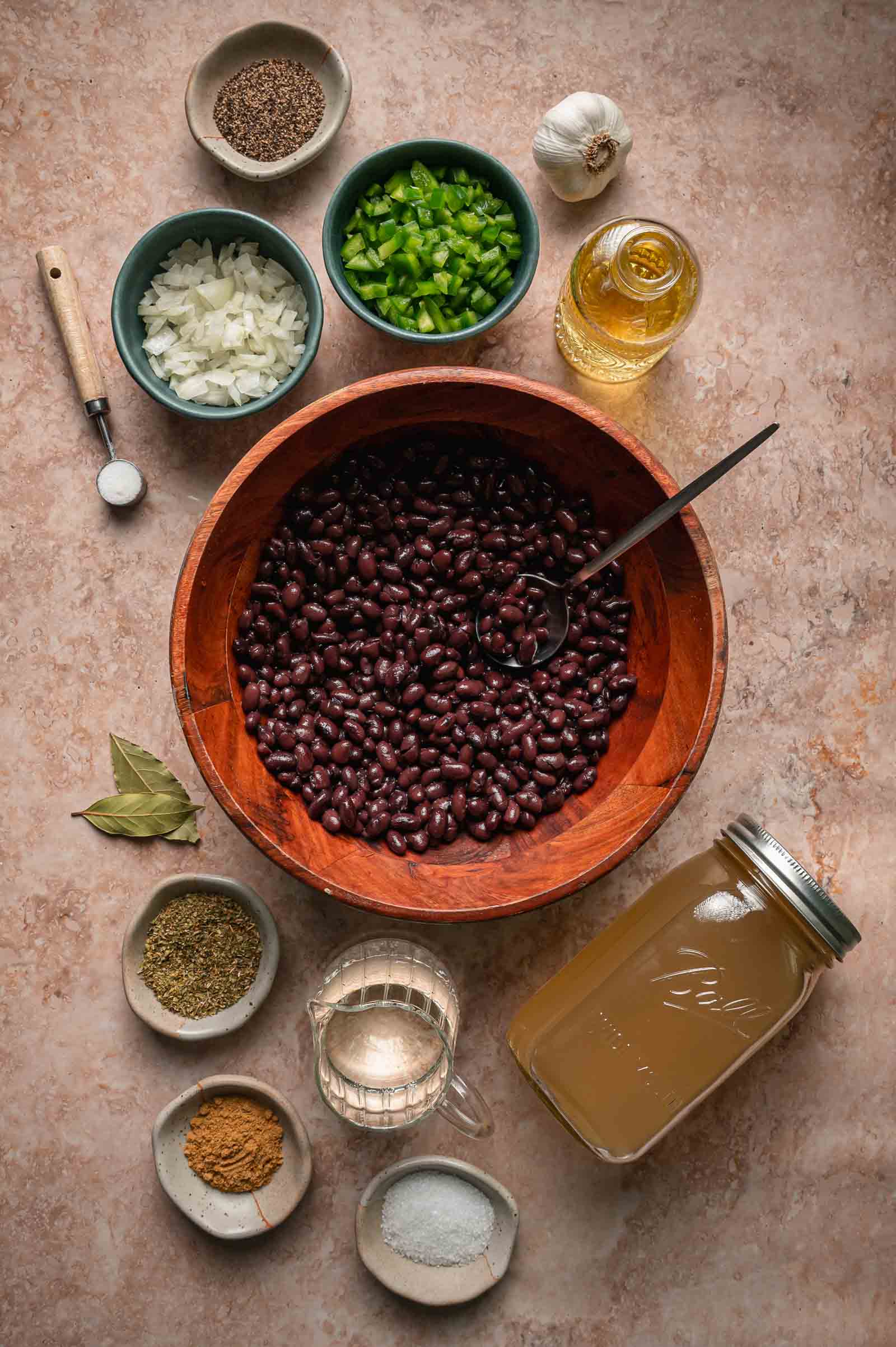 A bowl of black beans surrounded by chopped onions, peppers, spices, broth, garlic, oil, and salt on a countertop.