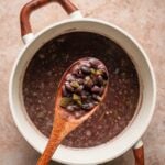 A wooden spoon holding cooked Cuban black beans and vegetables is held above a pot of black bean soup on a light brown surface.