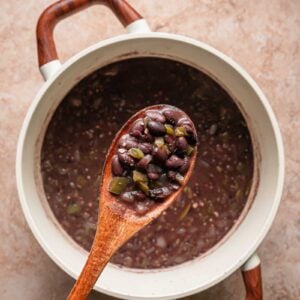 A wooden spoon holding cooked Cuban black beans and vegetables is held above a pot of black bean soup on a light brown surface.
