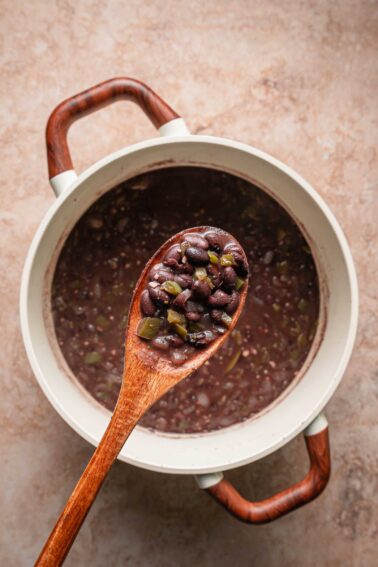 A wooden spoon holding cooked Cuban black beans and vegetables is held above a pot of black bean soup on a light brown surface.