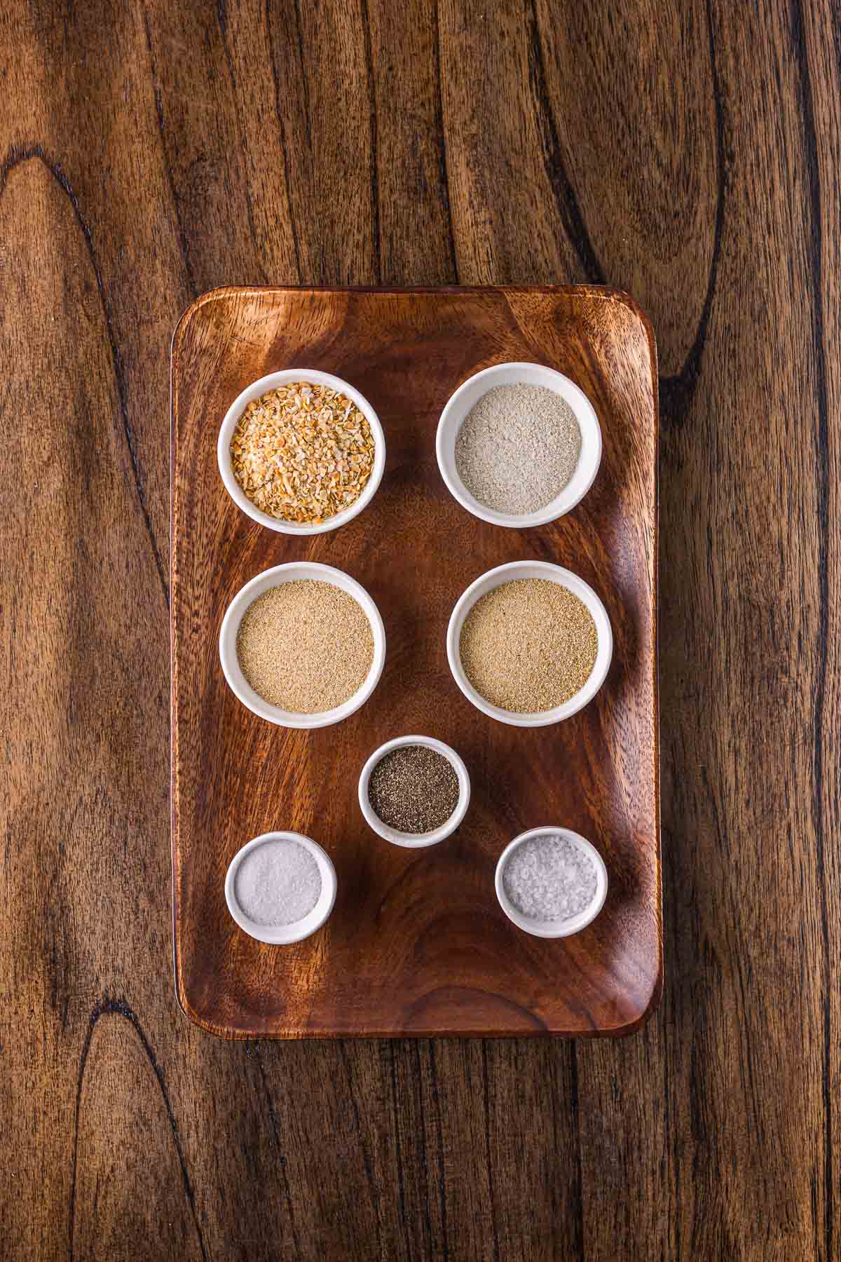 Seven small white bowls with various spices and seasonings are arranged on a rectangular wooden tray, set on a wooden surface.