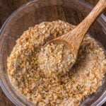 A clear glass bowl of french onion soup mix with a wooden spoon resting inside, on a wooden surface.