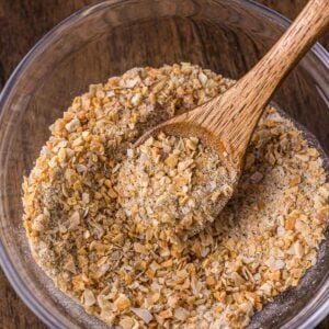 A clear glass bowl of french onion soup mix with a wooden spoon resting inside, on a wooden surface.