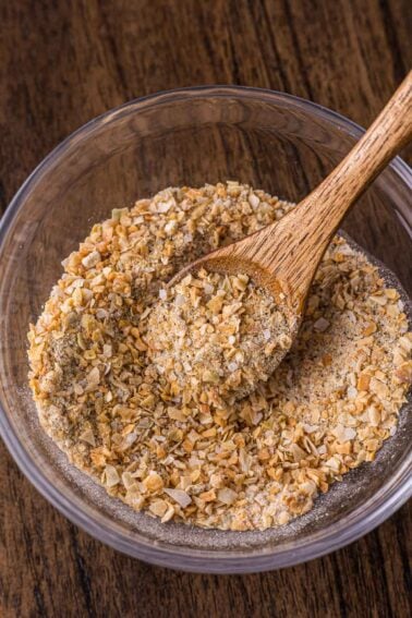A clear glass bowl of french onion soup mix with a wooden spoon resting inside, on a wooden surface.