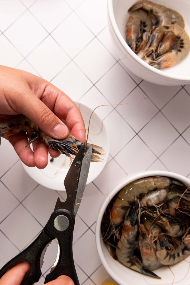 A hand uses kitchen shears to cut the legs off a raw shrimp over a white plate, with bowls of shrimp nearby on a tiled surface.