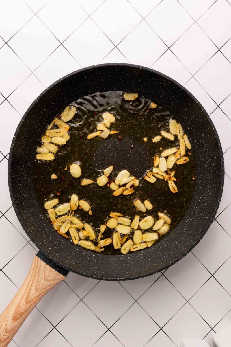 A black frying pan with sliced garlic frying in oil sits on a white tiled surface with a grid pattern.