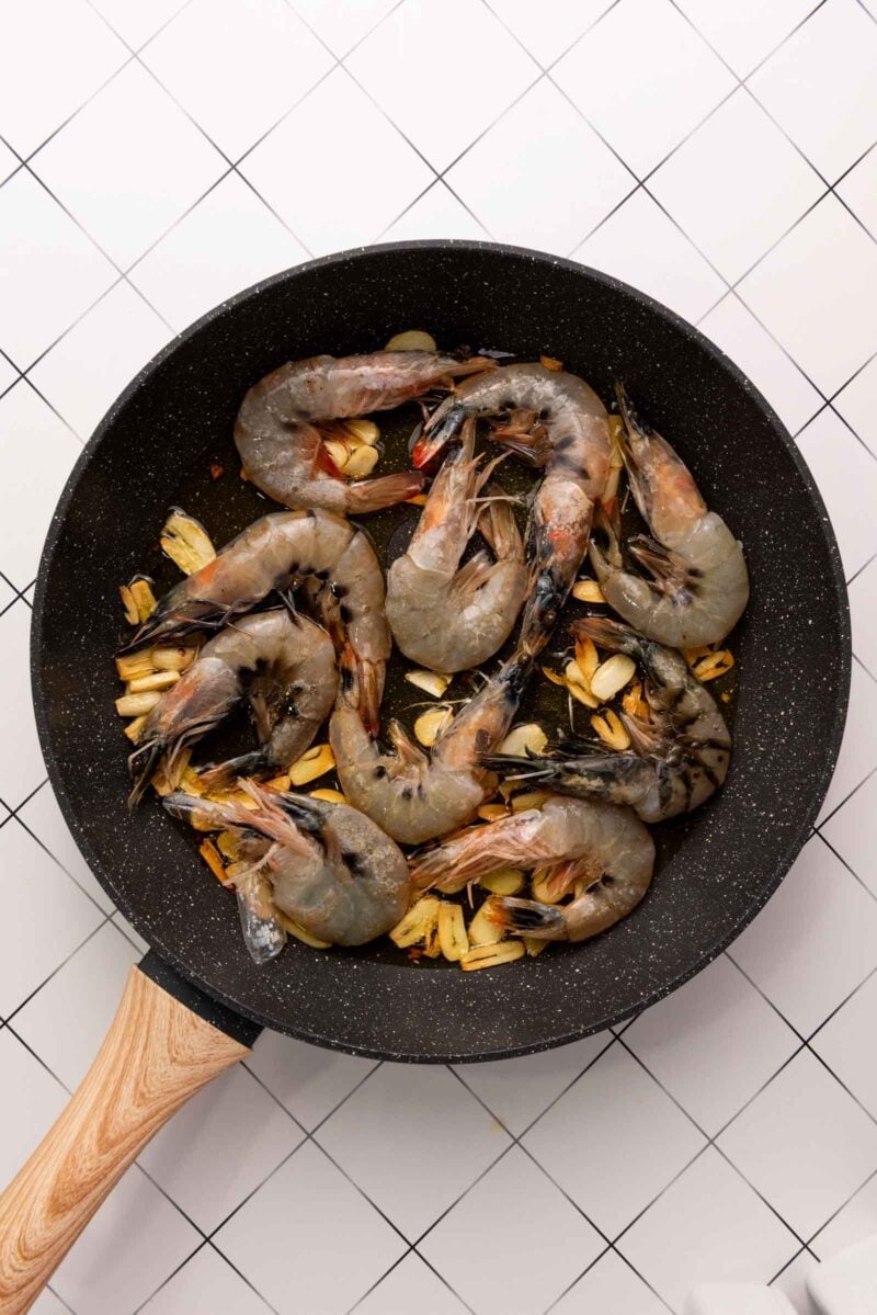 Raw shrimp and sliced garlic cooking in a black frying pan with a wooden handle, placed on a white tiled surface.