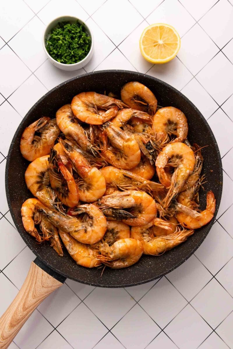 A skillet filled with cooked shrimp sits on a tiled surface, with a bowl of chopped herbs and a halved lemon nearby.