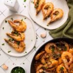 Cooked shrimp garnished with parsley on two white plates, with additional shrimp in a skillet, garlic cloves, and a green cloth nearby on a tiled surface.
