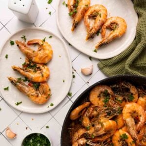 Cooked shrimp garnished with parsley on two white plates, with additional shrimp in a skillet, garlic cloves, and a green cloth nearby on a tiled surface.