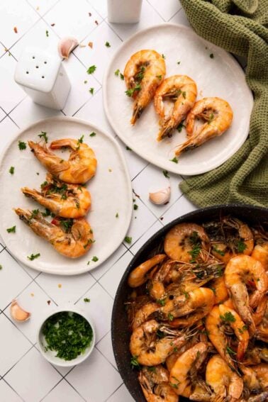 Cooked shrimp garnished with parsley on two white plates, with additional shrimp in a skillet, garlic cloves, and a green cloth nearby on a tiled surface.
