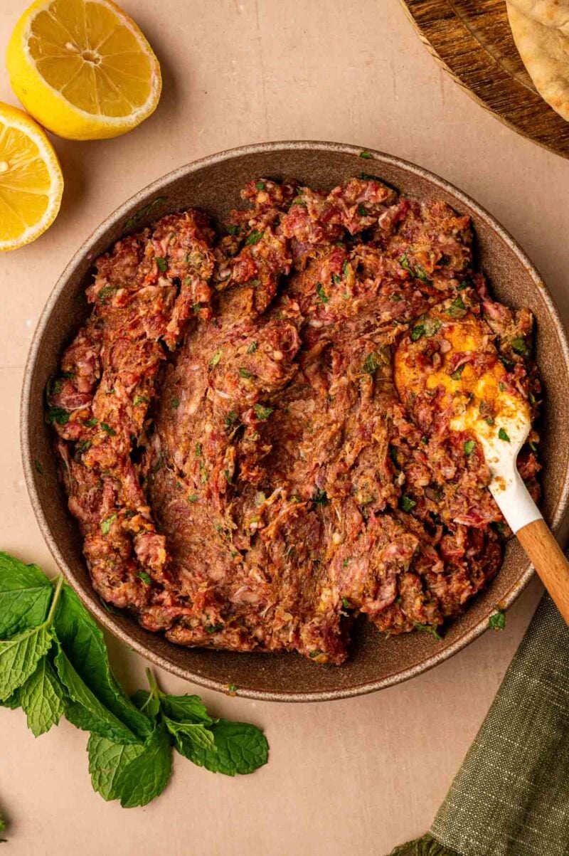 A bowl of spiced ground meat mixture with herbs, partially mixed with a spatula. Fresh mint, lemon halves, and flatbread are arranged nearby.