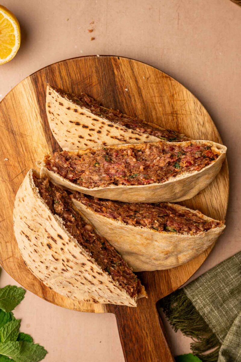 Three pita halves filled with a ground lamb and herb mixture are arranged on a round wooden board, with a lemon slice and mint leaves nearby.