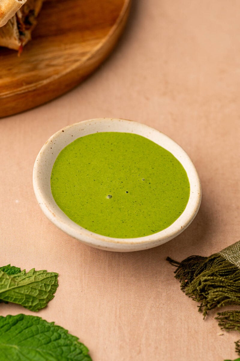 A small bowl of vibrant green sauce sits on a light-colored surface, with a few mint leaves and a green cloth nearby.