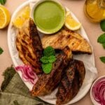 A plate with three grilled arayes, a bowl of green dipping sauce, lemon wedges, pickled red onions, and fresh mint, on a light surface with a green napkin.