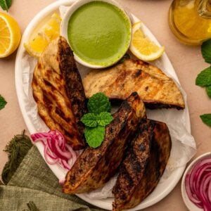 A plate with three grilled arayes, a bowl of green dipping sauce, lemon wedges, pickled red onions, and fresh mint, on a light surface with a green napkin.