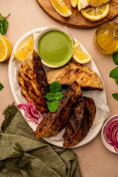A plate with three grilled arayes, a bowl of green dipping sauce, lemon wedges, pickled red onions, and fresh mint, on a light surface with a green napkin.