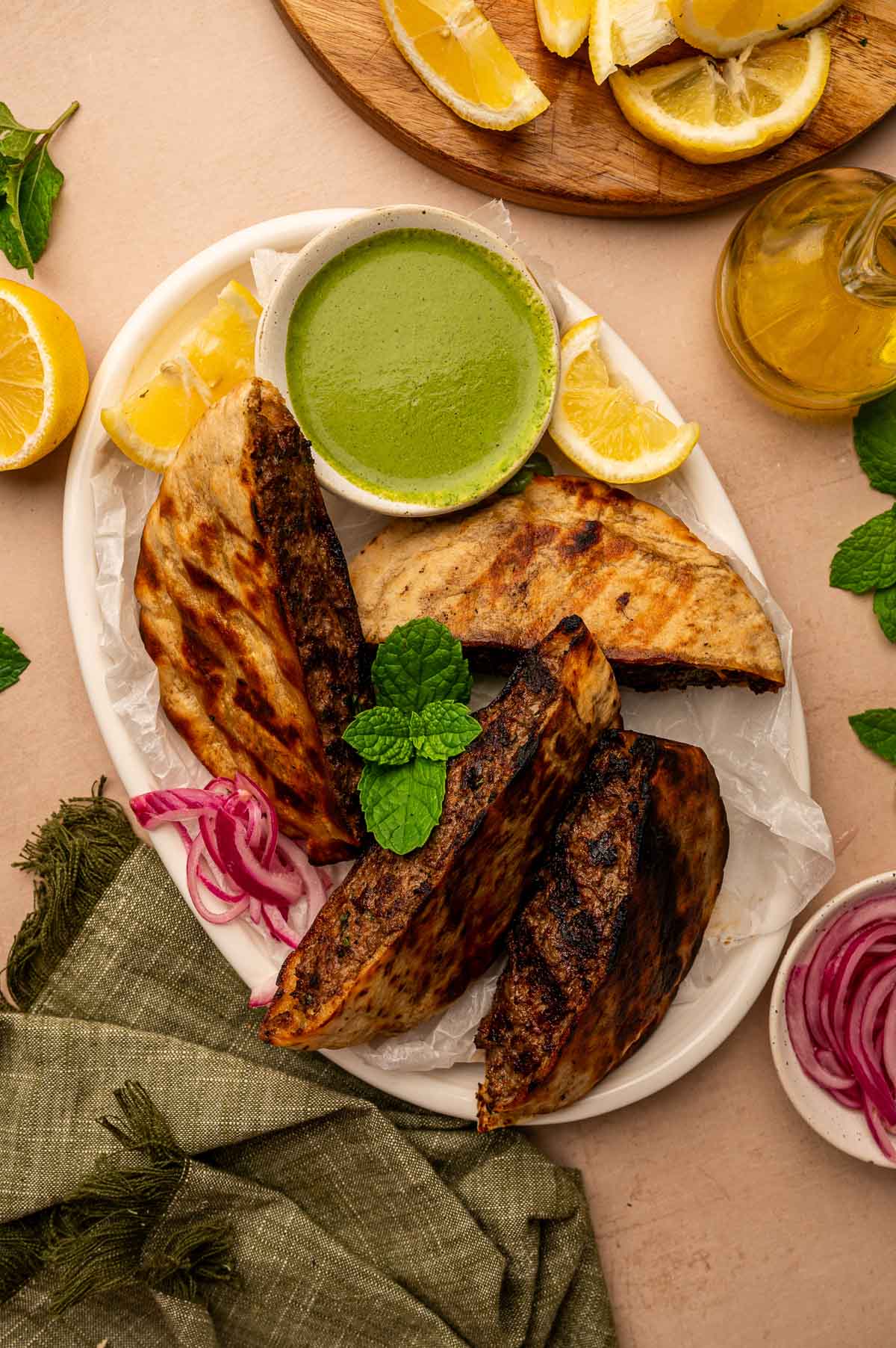 A plate with three grilled arayes, a bowl of green dipping sauce, lemon wedges, pickled red onions, and fresh mint, on a light surface with a green napkin.
